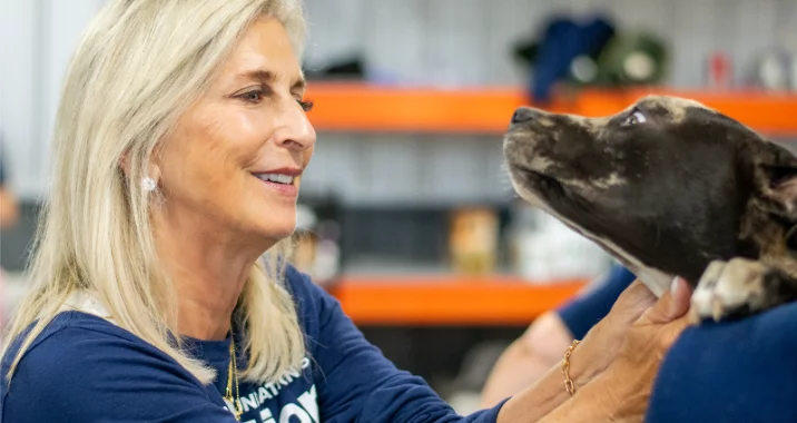 A smiling woman with blonde hair gently holds and looks at a black and white dog, making eye contact in an indoor setting with orange shelves in the background.