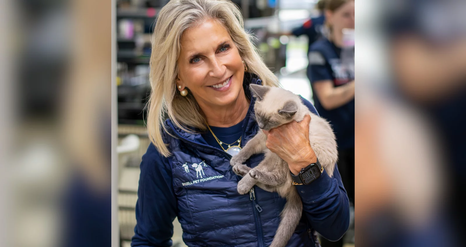 A smiling woman with long blonde hair holds a light brown kitten close to her chest. She is wearing a navy vest and standing indoors, with blurred people and shelves in the background.