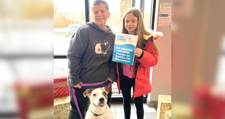A smiling adult and child pose indoors with a happy dog on a leash. The child holds a sign about pet adoption and supporting emptying shelters. Wrapped gifts are visible in the background.