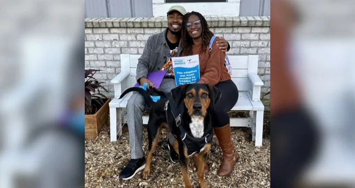 A smiling couple sits together on a white bench outside, holding a sign and leash. A large brown and black dog wearing a harness stands in front of them. There is a light brick wall and plants in the background.