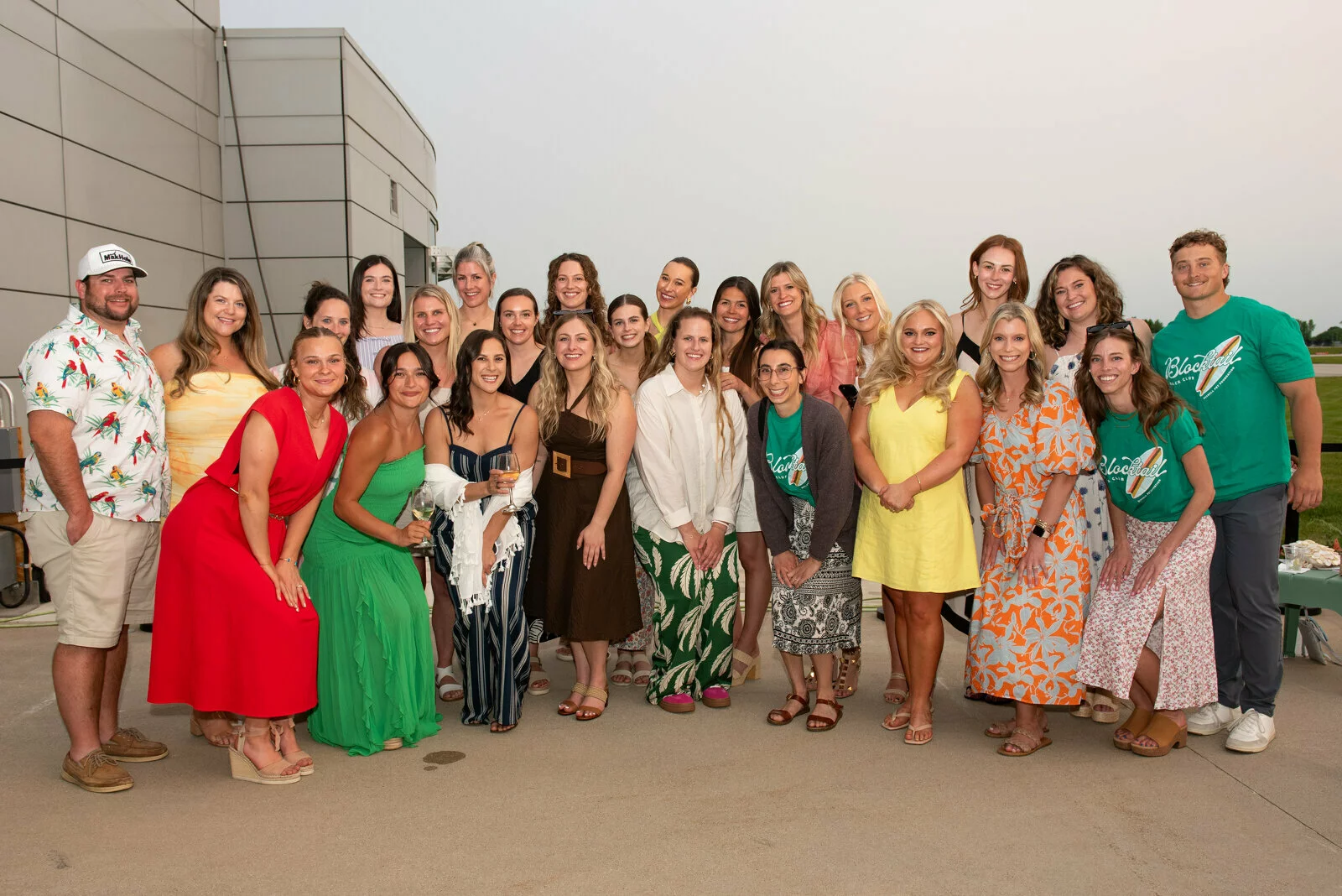 A group of 25 people, mostly women in colorful summer dresses, pose together and smile outdoors in front of a modern building on a clear day.