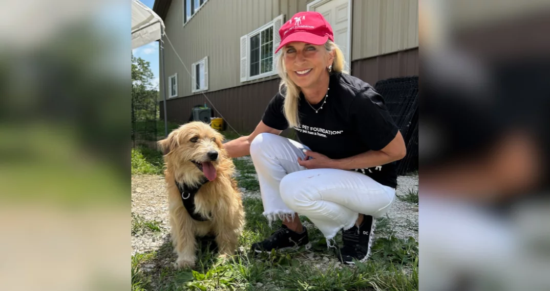 A smiling woman in a red hat and black shirt crouches next to a happy tan dog outdoors, with a light brown building and greenery in the background.