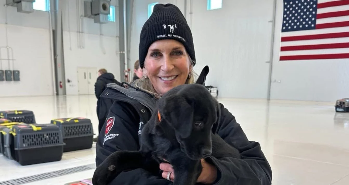 A smiling woman in a black beanie and jacket holds a black puppy in a large indoor space with an American flag on the wall and several pet carriers in the background.