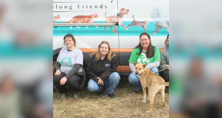 Three smiling women kneel on grass in front of an animal rescue van decorated with images of cats and dogs. A tan dog stands in the foreground, looking towards the camera.