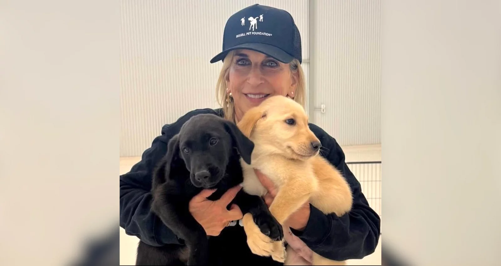 A smiling woman in a black cap and sweatshirt holds a black puppy in one arm and a yellow puppy in the other, standing in front of a light-colored, indoor background.