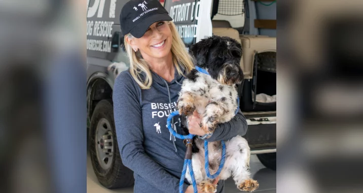 A smiling woman in a cap and hoodie holds a black and white dog on a blue leash, standing in front of a vehicle with dog rescue signage.