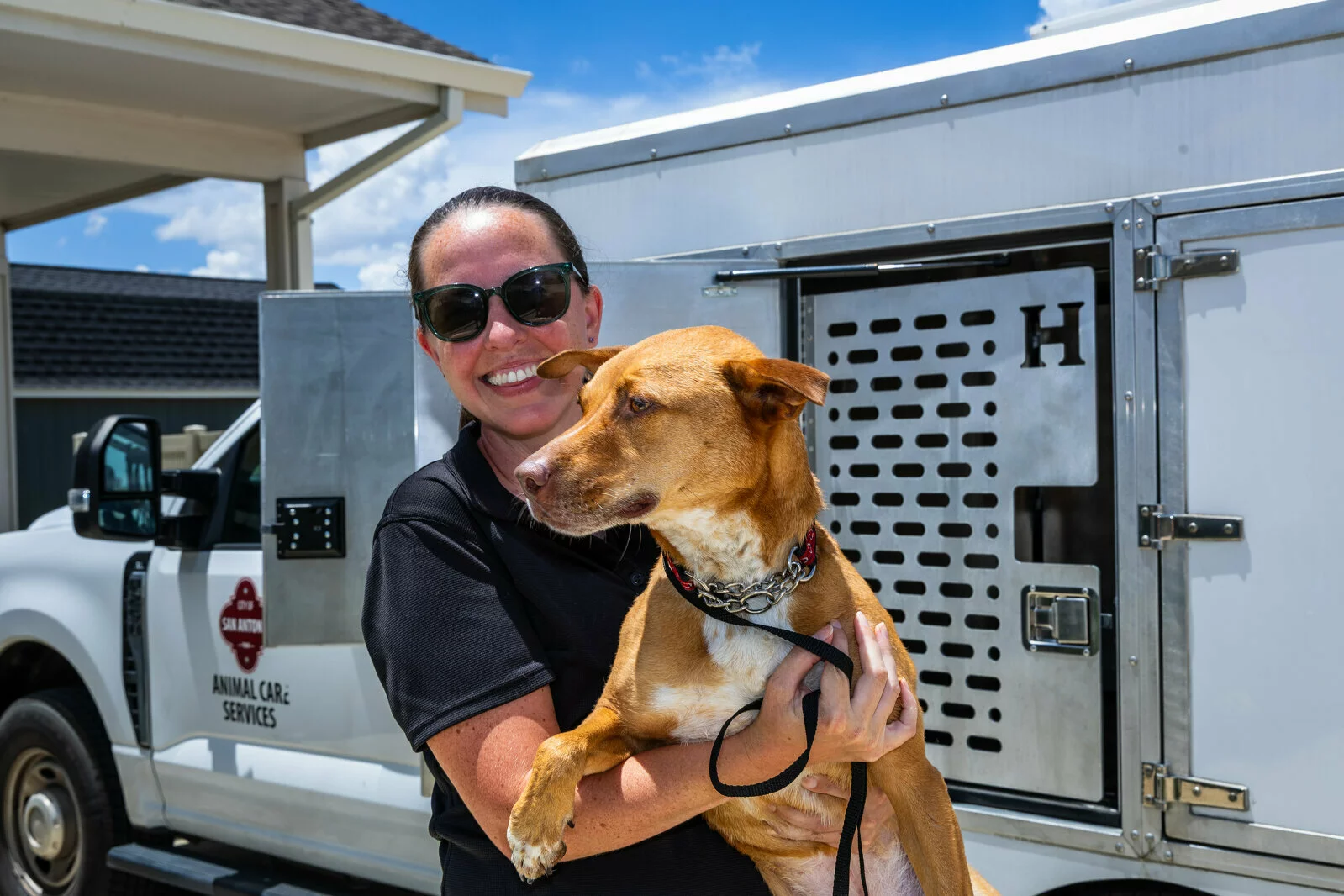 A smiling person in sunglasses holds a brown dog on a leash in front of an animal care services vehicle with a kennel compartment. The weather is sunny with a blue sky and some clouds.