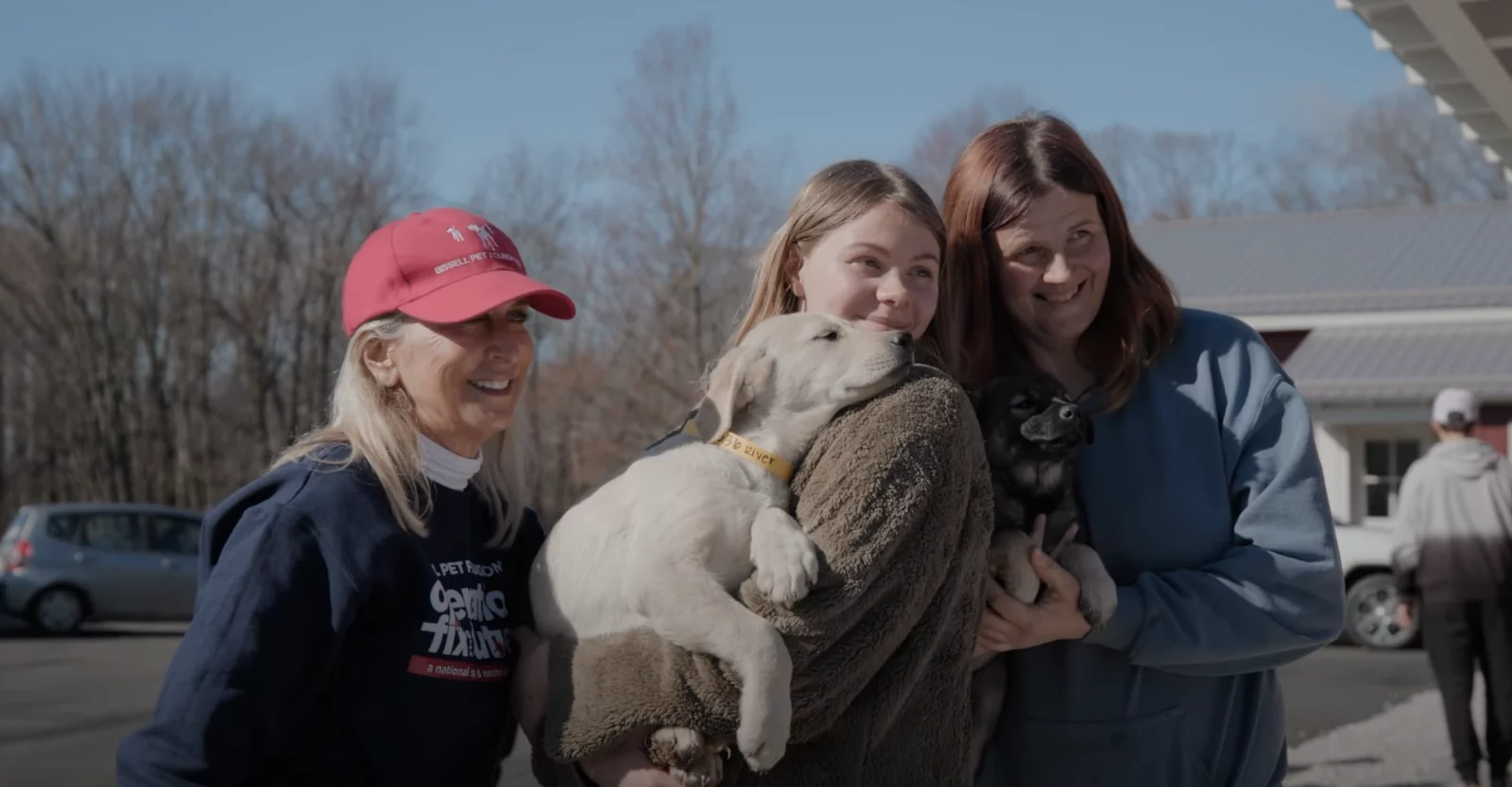Three women smile outdoors while holding two puppies, one light-colored and one dark, on a sunny day with bare trees and parked cars in the background.