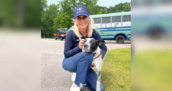 A woman in a navy hat and sweater crouches on grass, smiling and hugging a black and white dog. Behind them is a blue bus with dog images in the windows and trees in the background.