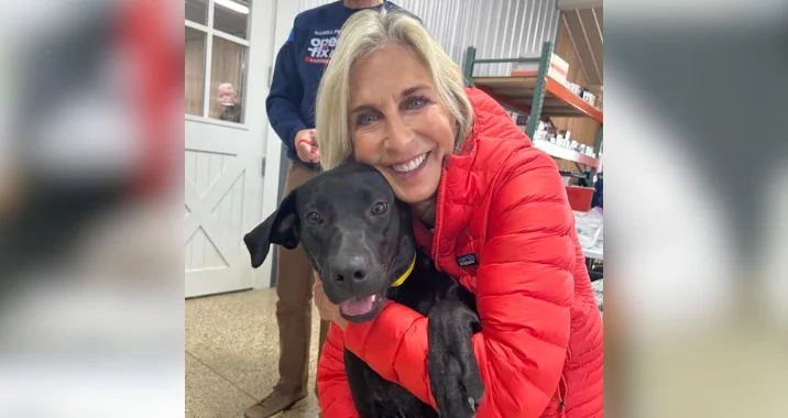 A smiling woman in a red jacket hugs a happy black dog indoors. The background shows another person standing and shelves with various items.